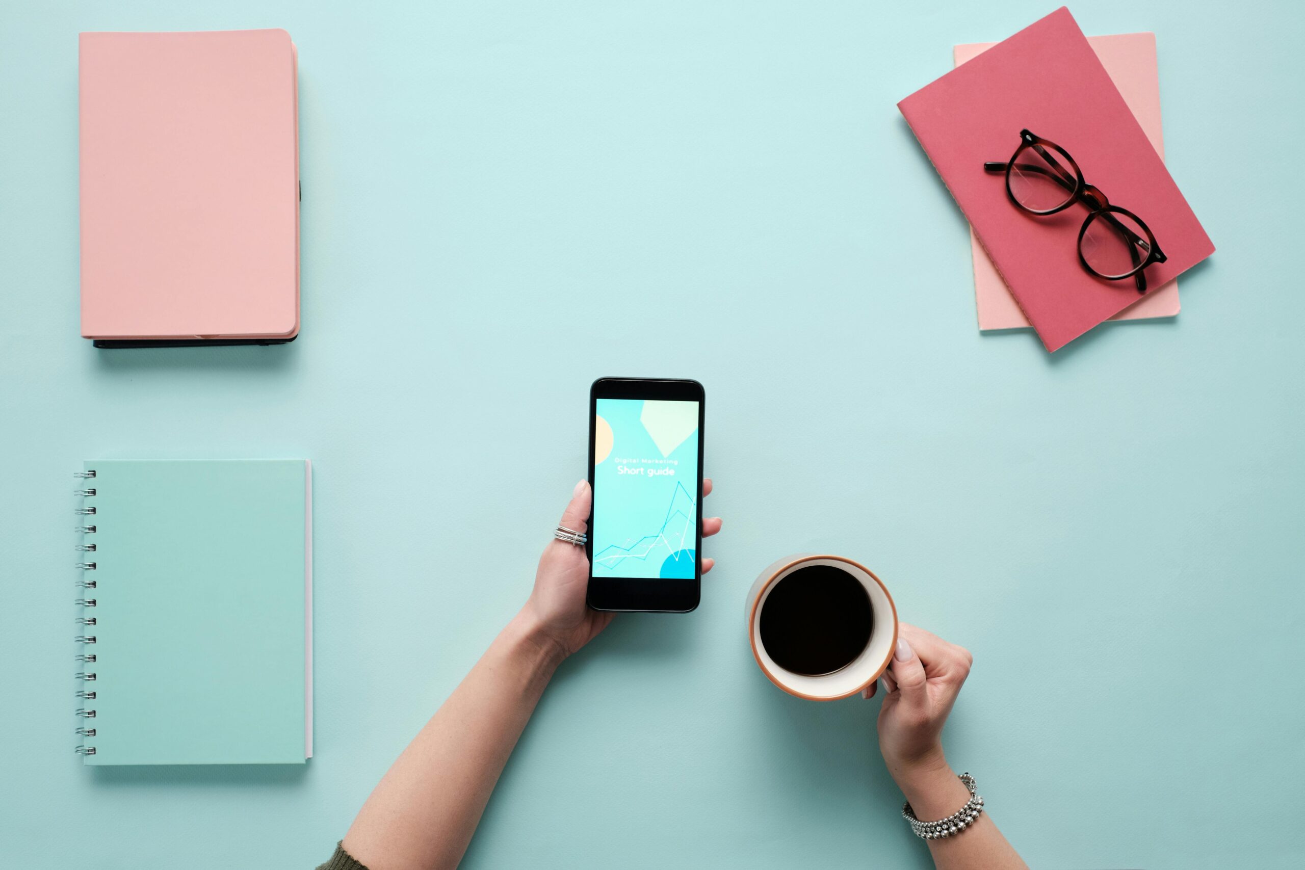 Overhead view of a stylish workspace featuring a smartphone, coffee, and notebooks on a pastel background.